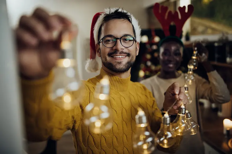 A man and woman hanging Christmas lights indoors, wearing festive hats and surrounded by holiday decorations.