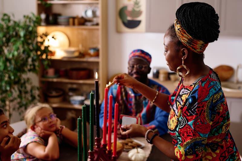 A mother and her family gathered around a table, lighting the kinara with food and decorations set for Kwanzaa.